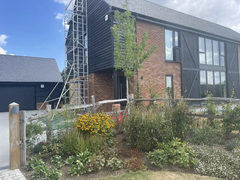 Modern barn style timber weatherboard home in Bossingham, near Canterbury, with newly planted borders in the foreground and a scaffold tower at the gable